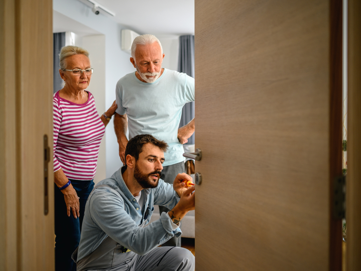 Man fixing a lock on the door while a senior couple is standing next to him.